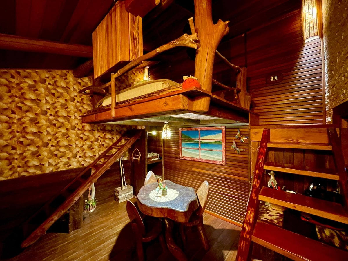 A suspended wooden bed is situated among tree branches, creating a unique sleeping area. Below, a small dining table is set with a floral centerpiece, surrounded by four chairs. Warm wooden walls and stairs lead to a cozy, rustic atmosphere.