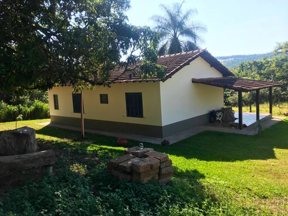 A charming rural house is set on a grassy area, featuring a porch supported by wooden posts. The exterior is painted in neutral tones, complemented by green window frames. Lush vegetation surrounds the property, contributing to the tranquil environment.