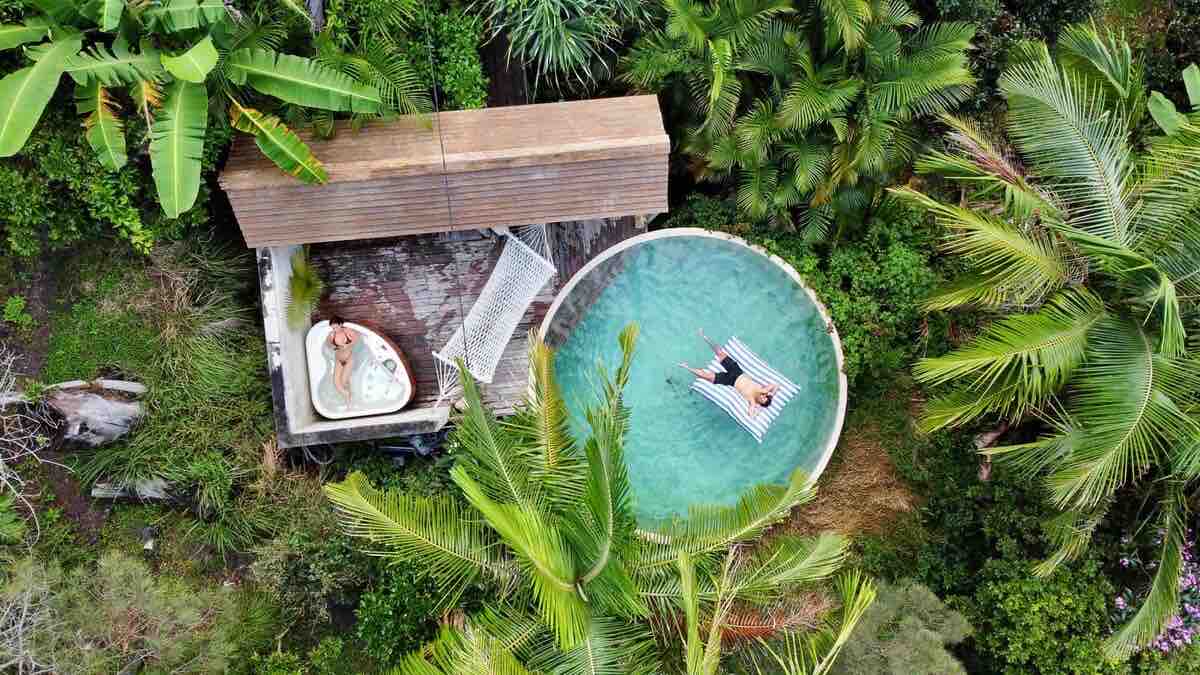 Aerial view captures a circular pool surrounded by lush tropical vegetation. A secluded jacuzzi is visible nearby, along with a shaded lounging area featuring comfortable seating. Palm trees frame the serene space, emphasizing the tranquil atmosphere of this private retreat.