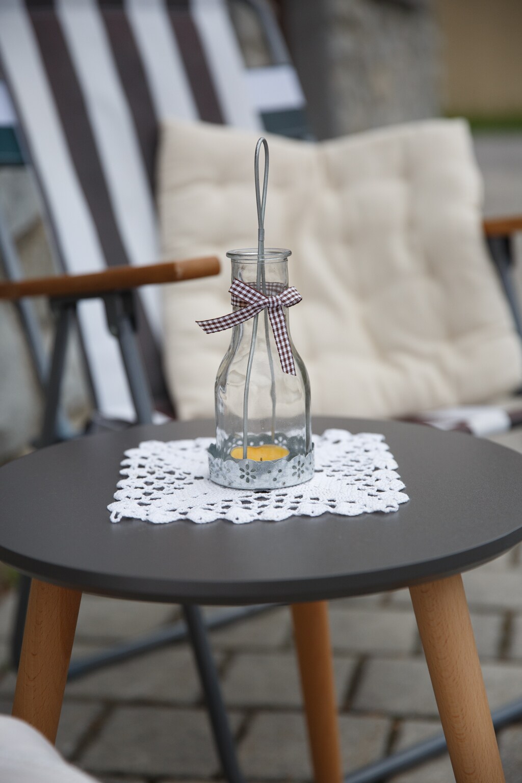 A small round table is positioned in front of two outdoor chairs. A glass bottle with a ribbon is placed on a lace doily at the center of the table. The scene conveys a sense of calm with natural tones and textures.