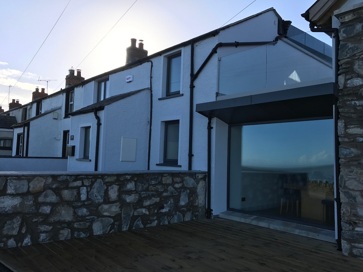 The exterior of a recently rebuilt cottage is displayed, featuring a combination of traditional stone and modern glass. The structure has multiple windows that allow natural light to shine in, while a wooden deck enhances the outdoor space, with views of the surrounding area visible.