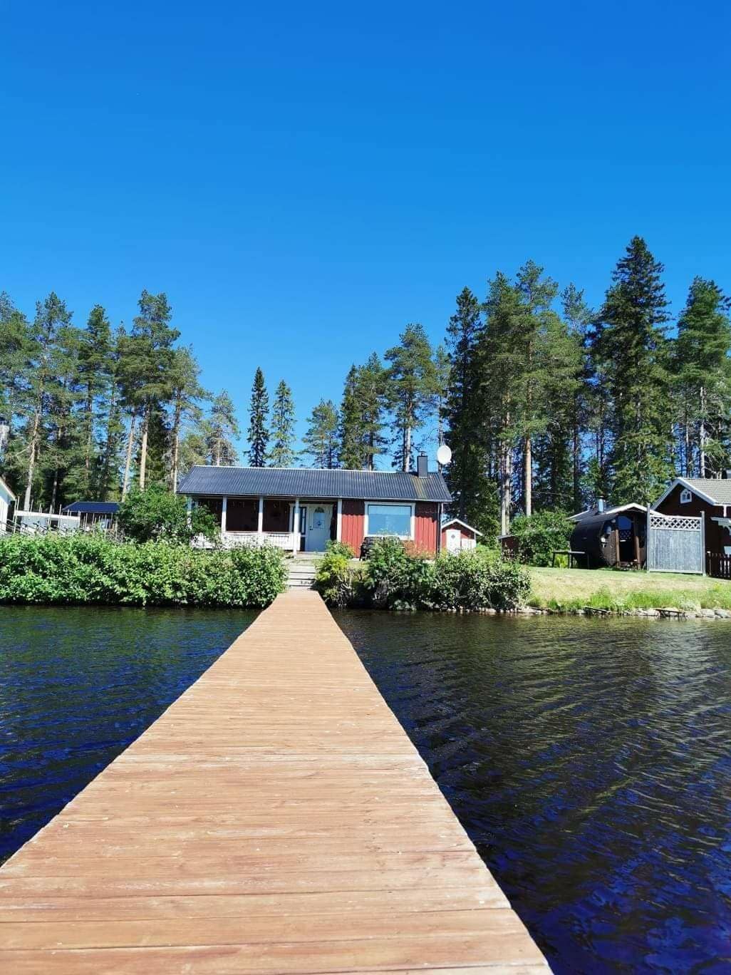 A wooden pier extends toward a charming house with a red facade, surrounded by a vibrant green landscape. Tall trees line the background, and the clear water reflects the blue sky, creating a serene view of the water's edge.