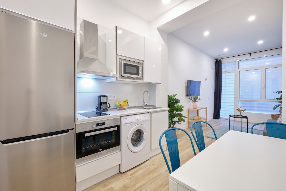 A modern kitchen area features stainless steel appliances, including a refrigerator and oven. A washer is included for convenience. A white dining table is surrounded by transparent blue chairs. Natural light is provided by large windows in the background, highlighting indoor plants.