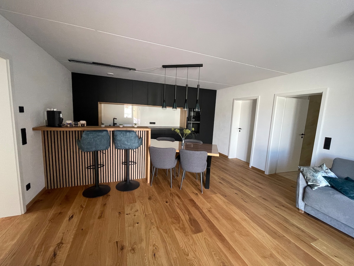 The dining area features a dark wood table surrounded by four chairs, complemented by two bar stools at a breakfast bar. A sleek kitchen with modern cabinetry is visible in the background, harmonizing with the warm wooden flooring and bright wall colors.