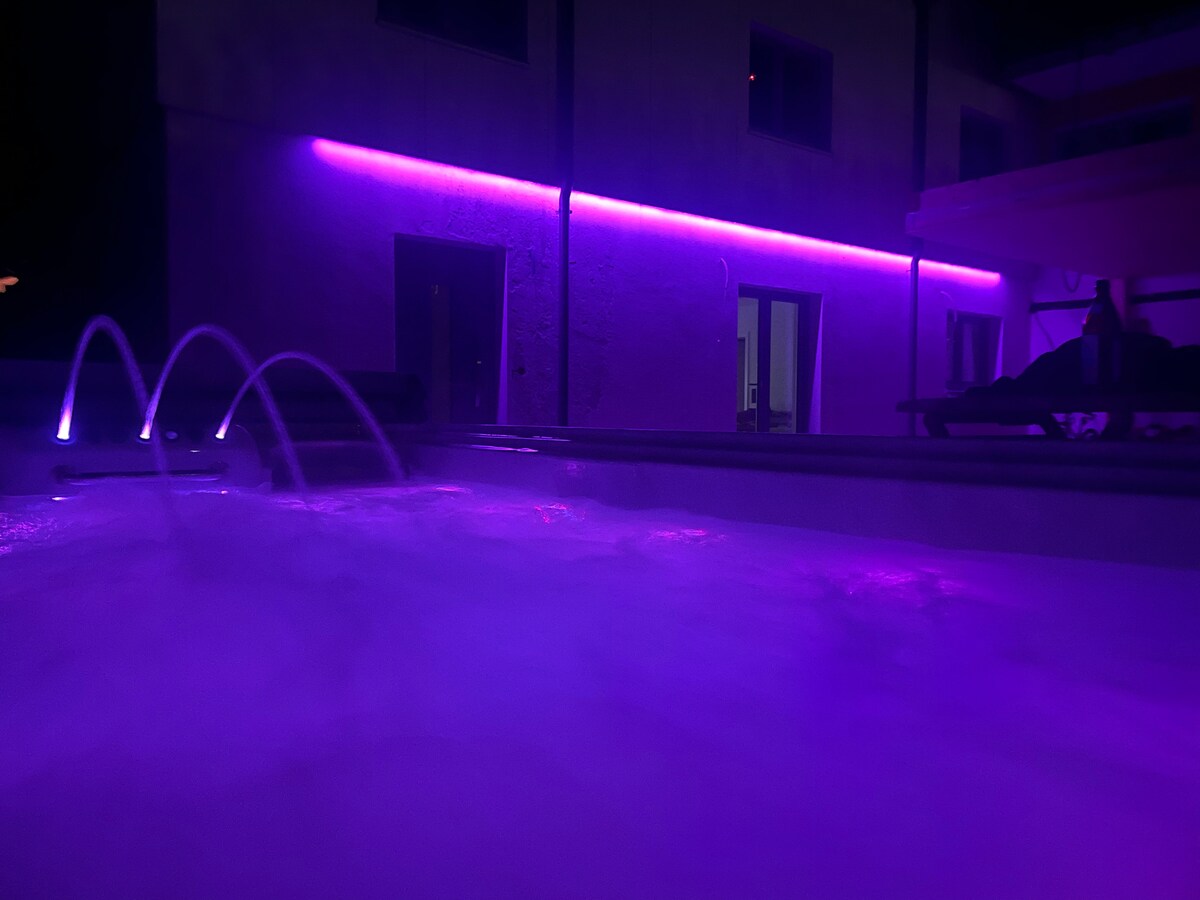 A swim spa is illuminated with soft purple lighting, surrounded by a misty atmosphere. Water gently cascades from spouts into the spa, creating a tranquil setting at night. The background showcases doors leading to the interior, softly reflecting the ambient glow.