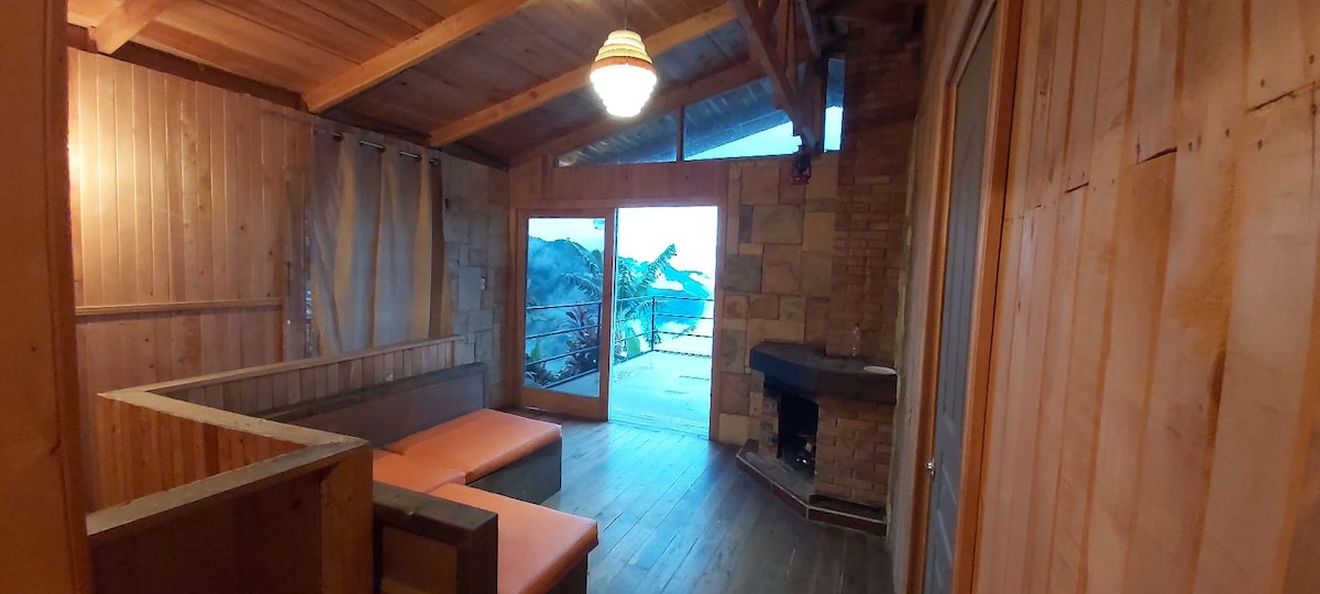 The inviting living area features natural wooden walls and a comfortable seating arrangement. A large glass door opens to a scenic outdoor view, allowing natural light into the space. A stone fireplace is present, contributing to a cozy ambiance.