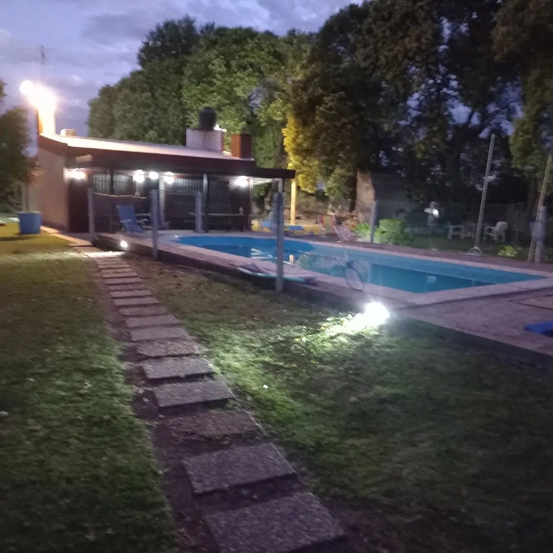 The outdoor area features a swimming pool, illuminated by soft lighting, surrounded by a stone pathway and well-maintained grass. In the background, a patio with seating is visible under trees, creating a serene setting for relaxation.