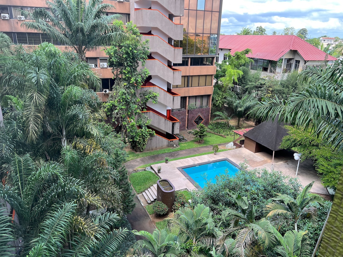 A view of the outdoor area features lush green foliage surrounding a serene pool area. The pool is framed by manicured lawns and a gazebo, with multiple buildings visible in the background, showcasing a mix of modern architecture and landscaping.