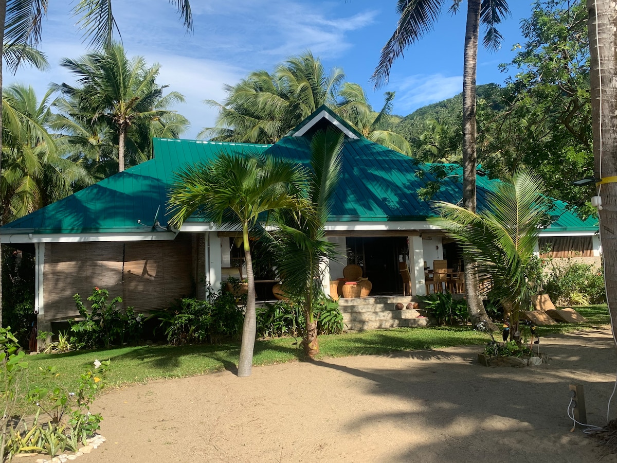 A spacious beach house is surrounded by tropical greenery, featuring palm trees and well-maintained gardens. The structure showcases a green roof and a large terrace. Sunlight dapples the sandy area in front, creating a relaxed outdoor setting with ample space for seating.