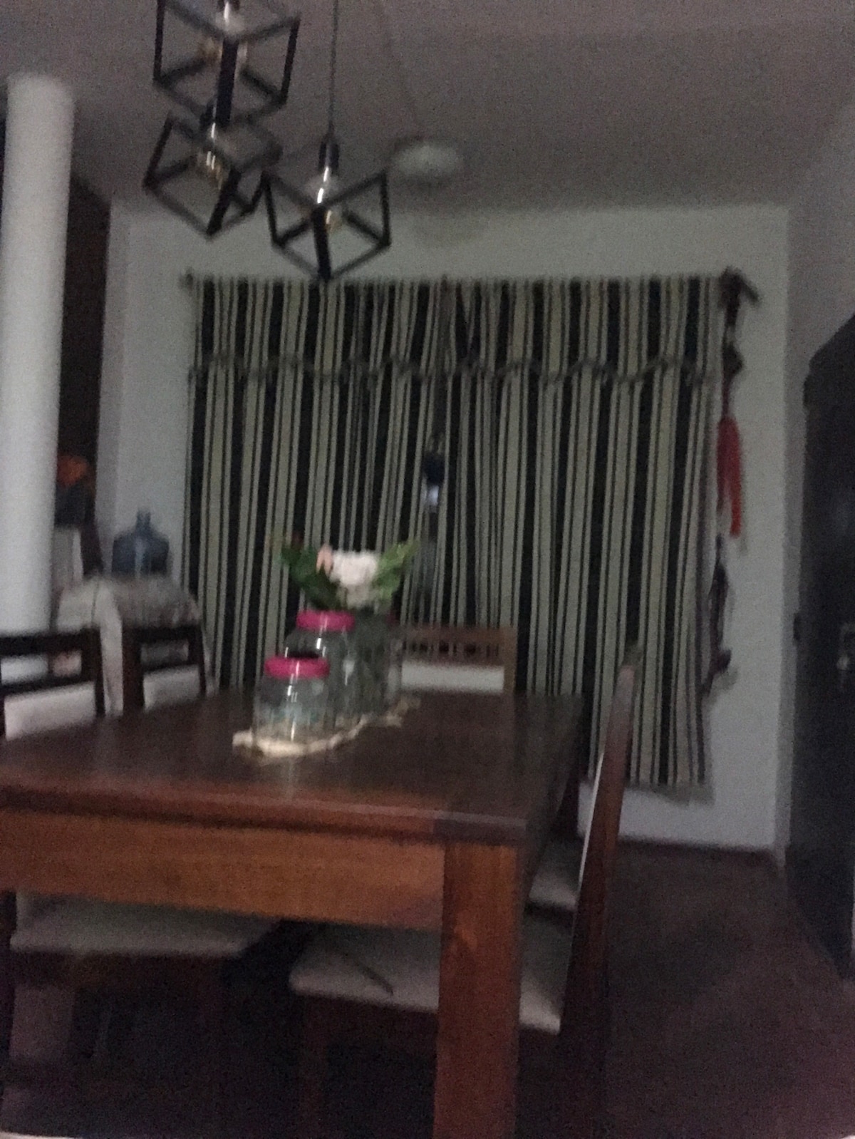 A dining area features a wooden table surrounded by several chairs, with a decorative table runner. A bouquet of flowers is placed in a glass vase at the center. Striped curtains frame a window, and geometric light fixtures hang above the table.