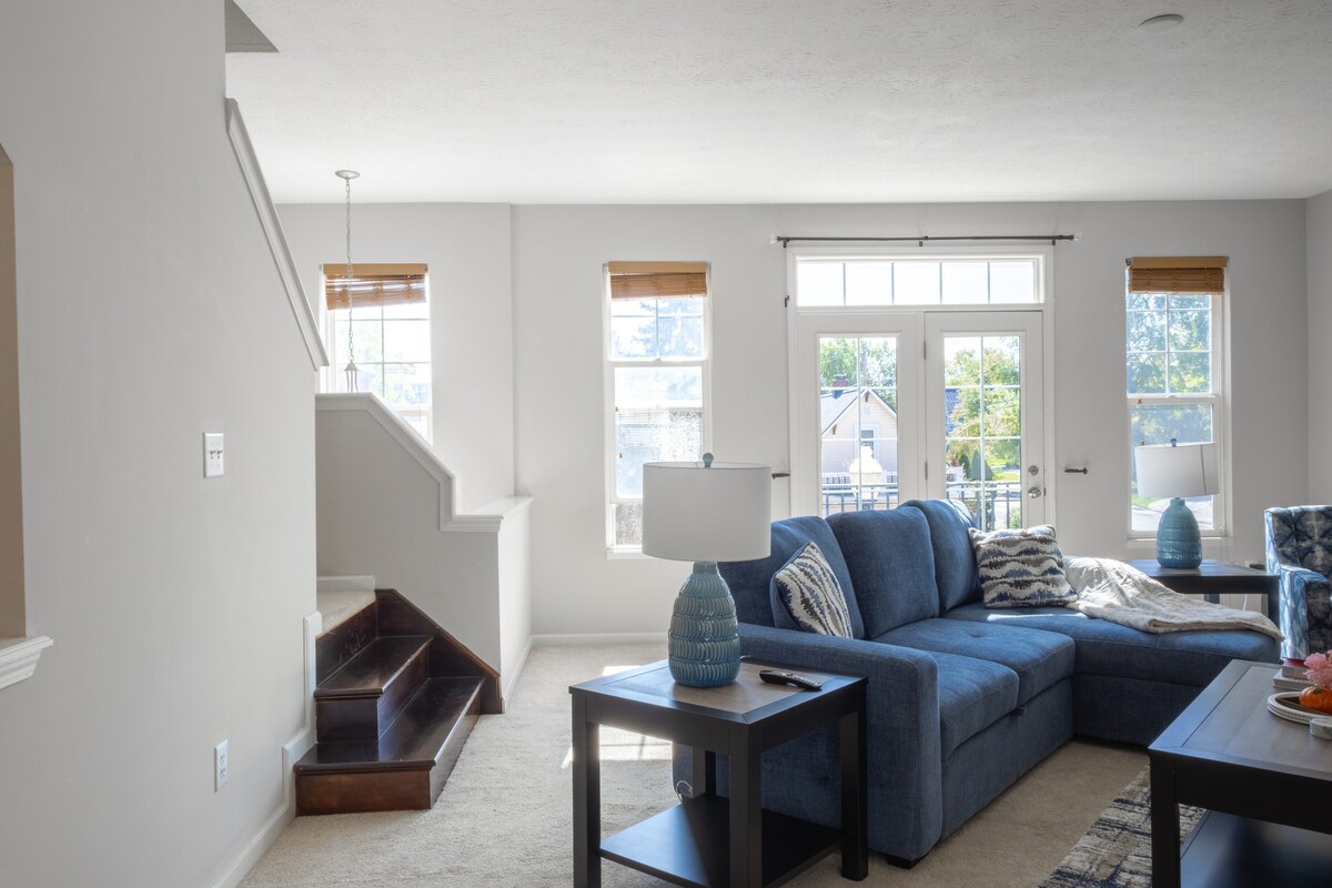 A spacious living area is highlighted by a blue sectional sofa, paired with a wooden coffee table. Natural light filters through two sets of windows, while light-colored walls enhance the bright atmosphere. A staircase is visible in the background, featuring a natural wood finish.