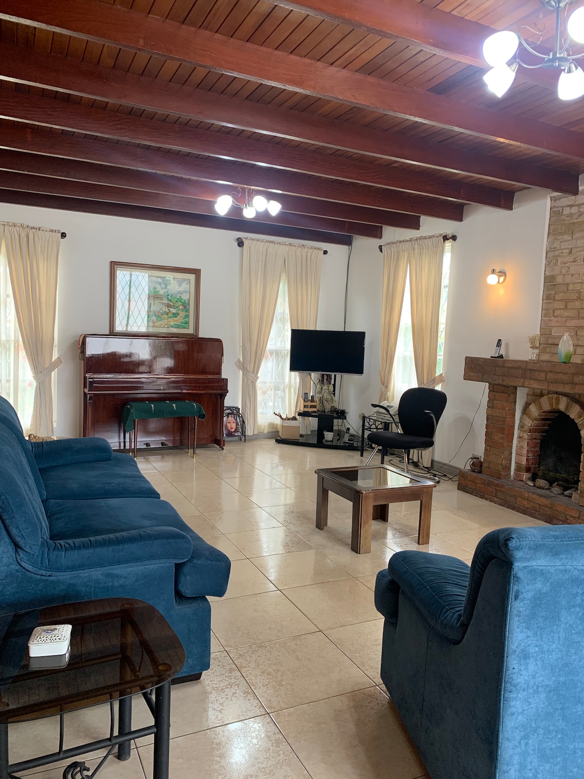 A spacious living room showcases high wooden beams and natural light filtering through sheer curtains. Comfortable blue sofas are arranged around a low coffee table, with a flat-screen television and a piano visible. A rustic fireplace adds a cozy element to the room.
