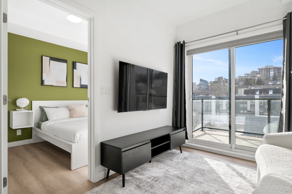 A bright living area is highlighted by a large wall-mounted TV and a comfortable couch. A sliding glass door leads to a balcony, offering views of the city. The adjoining bedroom features a spacious bed and a fresh green accent wall, creating a modern atmosphere.