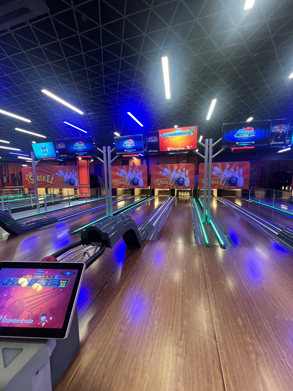 A modern bowling alley features several lanes equipped with glowing neon lights and electronic score displays. Polished wooden floors reflect the ambient lighting, while multiple screens showcase game information. The overall layout indicates a vibrant and engaging atmosphere for leisure activities.