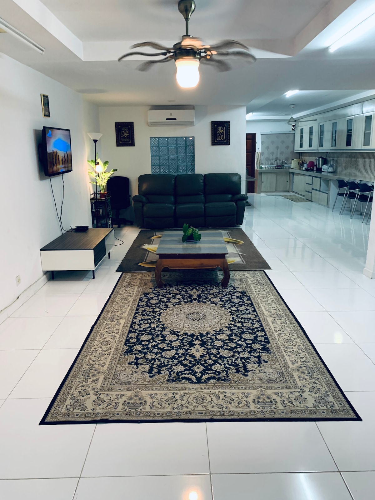 A spacious living area is furnished with a dark leather sofa, arranged around a central coffee table on a patterned rug. A television is mounted on the wall, and a large kitchen area is visible in the background, featuring modern cabinetry and seating.