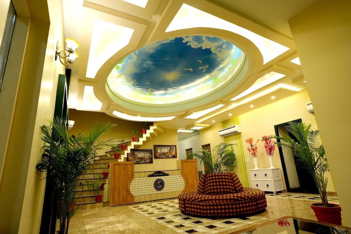 The interior entrance features an expansive, decorative ceiling painted with a serene sky and birds. Lush green plants frame the space, and a cozy, checkered seating area invites relaxation. A staircase leads to additional areas, illuminated by soft, ambient lighting.