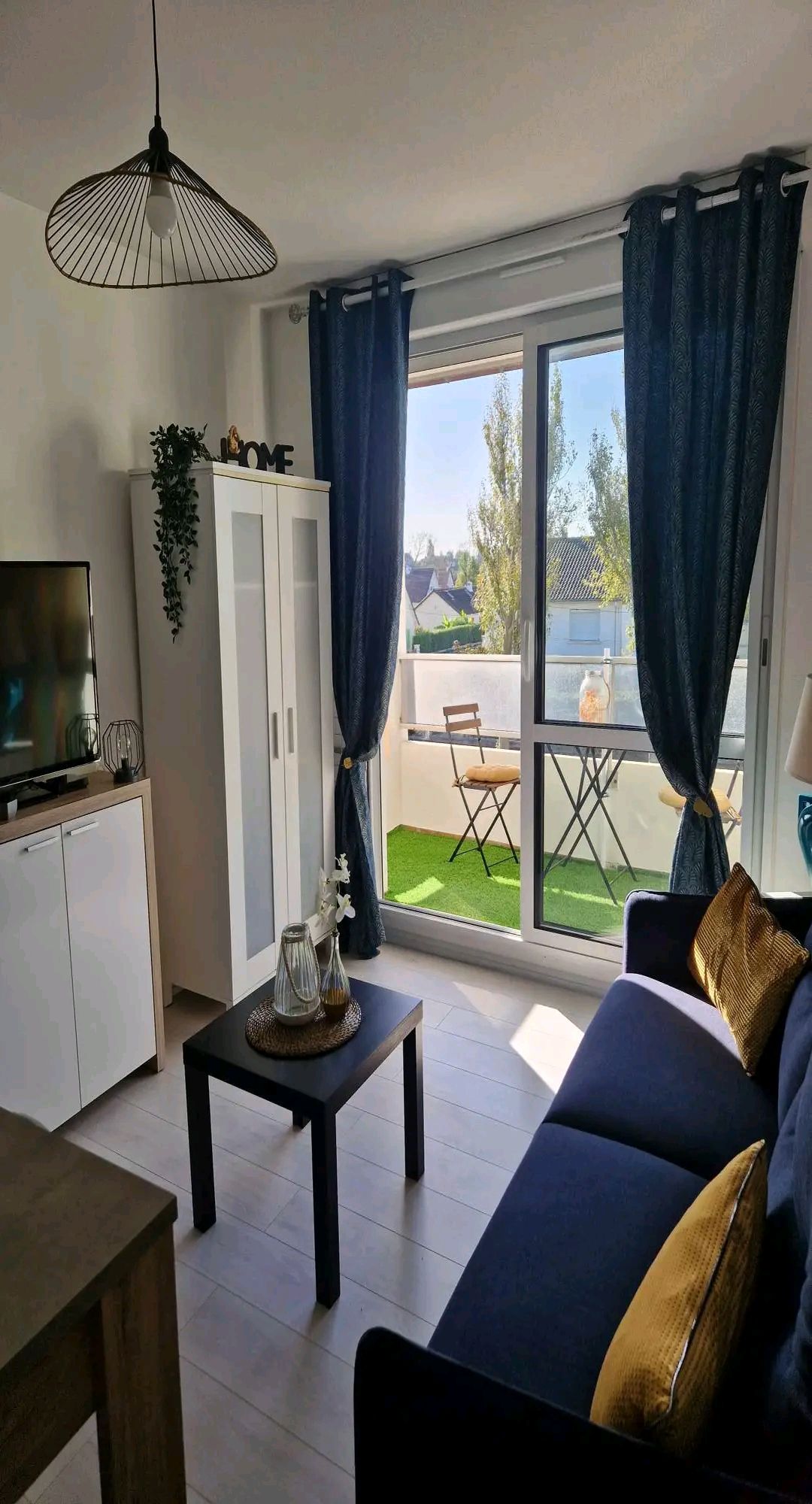 A light-filled living area features a navy blue sofa adorned with decorative cushions. A small coffee table sits in front of the sofa. A cabinet stands against the wall, while large glass doors open to a balcony with two chairs and a table, set against greenery.