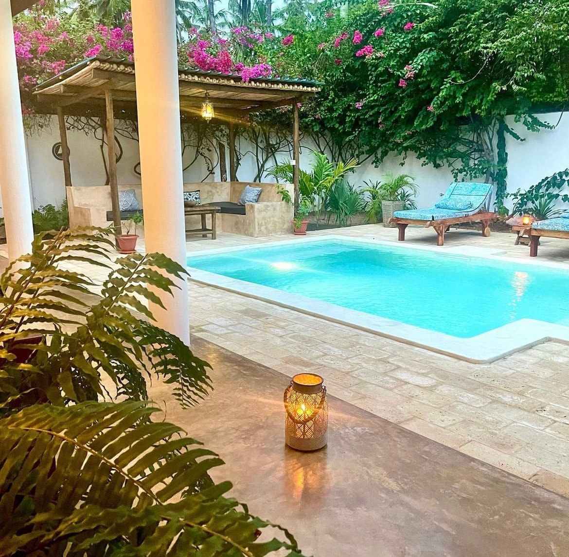 A serene outdoor space features a small private pool surrounded by lush greenery and flowering plants. Comfortable sun loungers and a shaded seating area provide relaxation options. Soft lighting from decorative lanterns enhances the ambiance during the evening hours.
