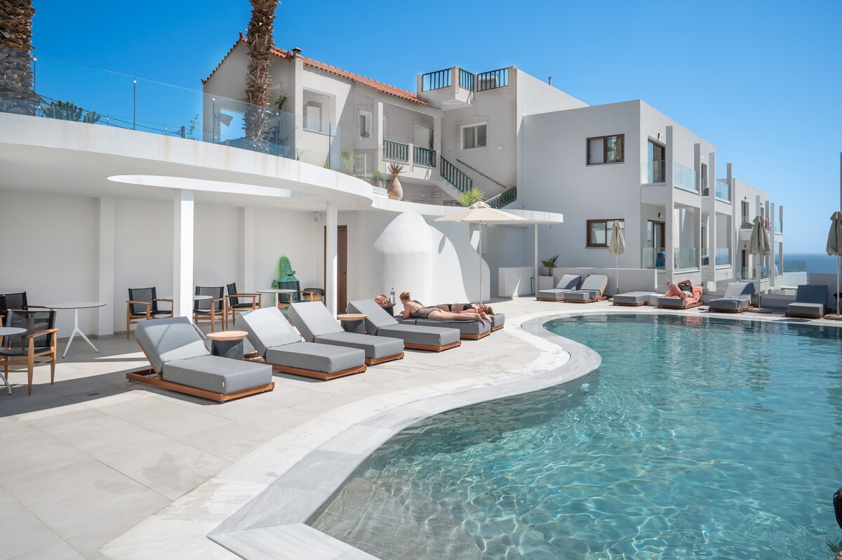 A shared outdoor swimming pool is surrounded by lounge chairs and a sun terrace, with sunbathers relaxing under clear blue skies. The modern architecture features white walls and an array of palm trees in the background, enhancing the inviting environment.