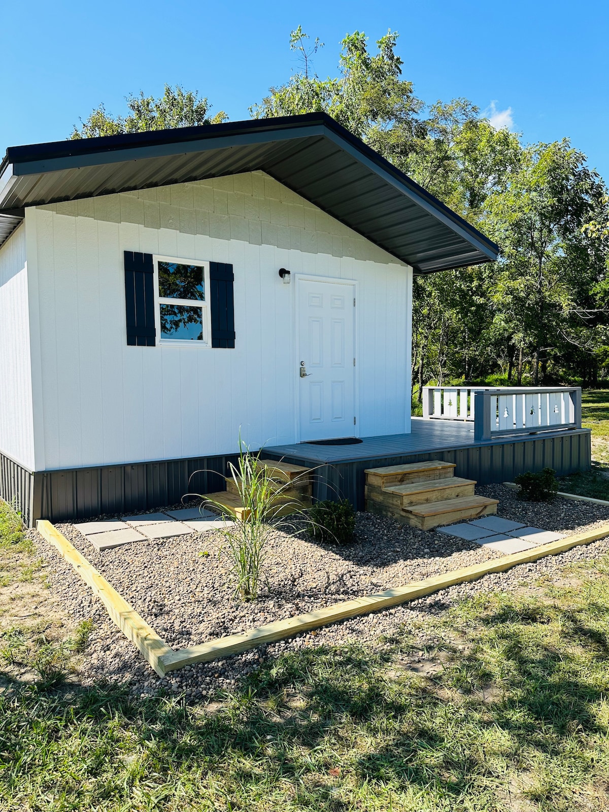 A small cottage with a sloped roof is presented, featuring white paneling and dark blue trim. A wooden porch with steps is visible, along with a neatly landscaped area of stones and grass surrounding the cottage. Bright blue skies and trees are seen in the background.