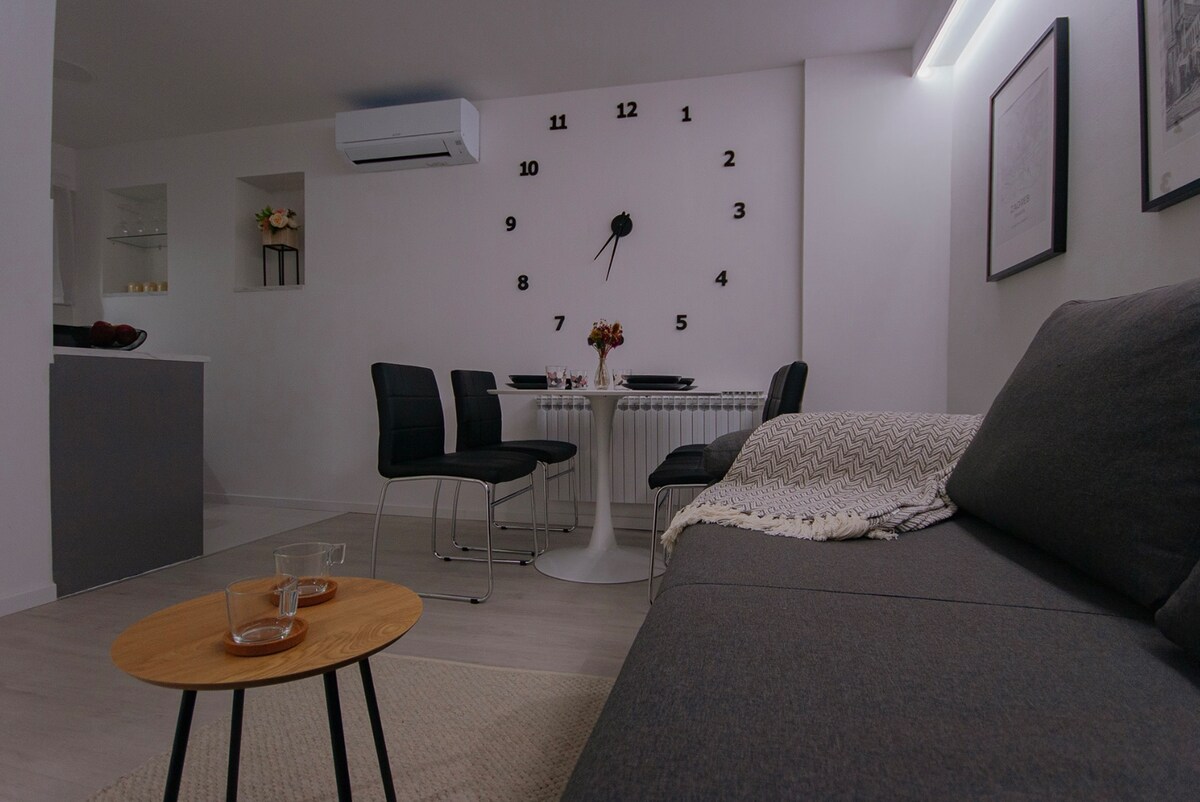 A modern living space is presented with a comfortable gray sofa and a stylish round dining table set for two, complemented by black dining chairs. A prominent wall clock adds a functional decor touch, while soft lighting enhances the overall ambiance.
