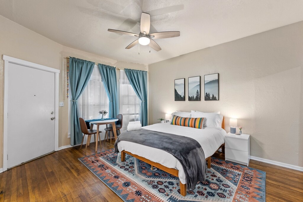 A spacious bedroom features a queen bed dressed in crisp linens, complemented by decorative pillows. Natural light filters through large windows, adorned with teal curtains. A small dining table with two chairs is situated near the entrance, while the ceiling fan provides a cooling breeze.