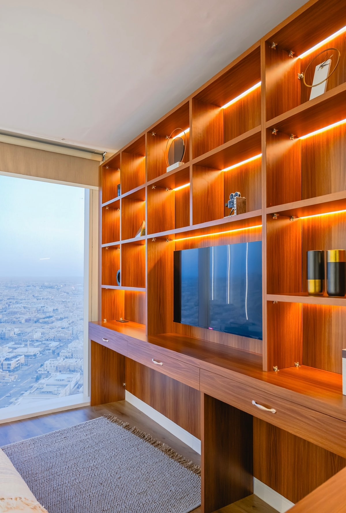 A modern wooden shelving unit features integrated lighting, providing a warm glow. A television is mounted on the wall, while decorative items and vases are displayed. A large window offers views of the city skyline beyond.