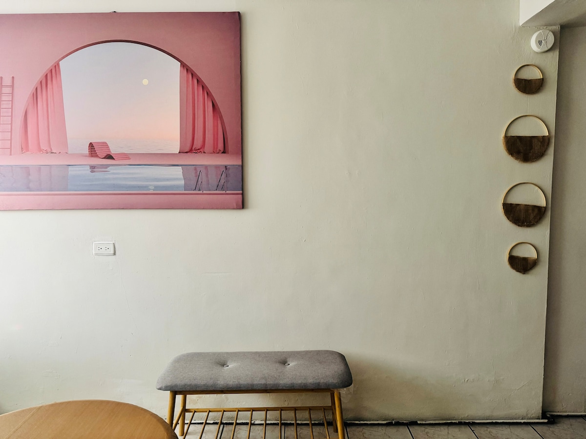 A colorful artwork featuring a sunset scene is displayed on the wall, complemented by decorative wooden baskets hanging above. A simple gray bench sits below the artwork, while a wooden table is partially visible to the left.