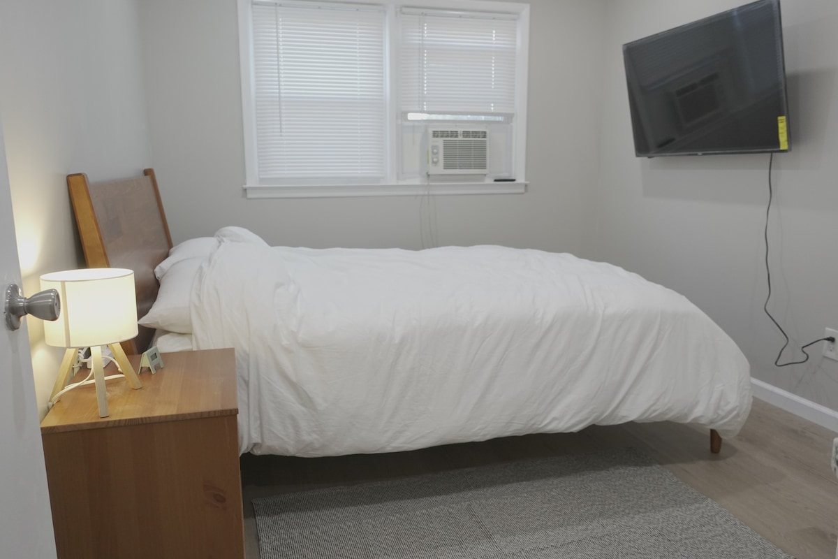 A cozy private room features a spacious bed with a white comforter, complemented by a wooden nightstand with a lamp. Natural light enters through two windows, and a wall-mounted TV is present opposite the bed. An air conditioning unit is installed for comfort.