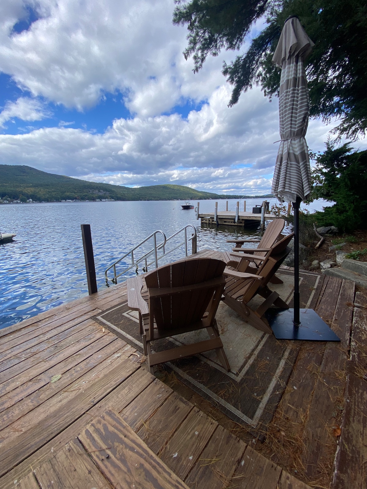 A wooden dock extends into the lake, featuring two Adirondack chairs and a large umbrella providing shade. The water reflects a mix of blue sky and clouds, while mountains frame the background, creating a serene lakeside environment.
