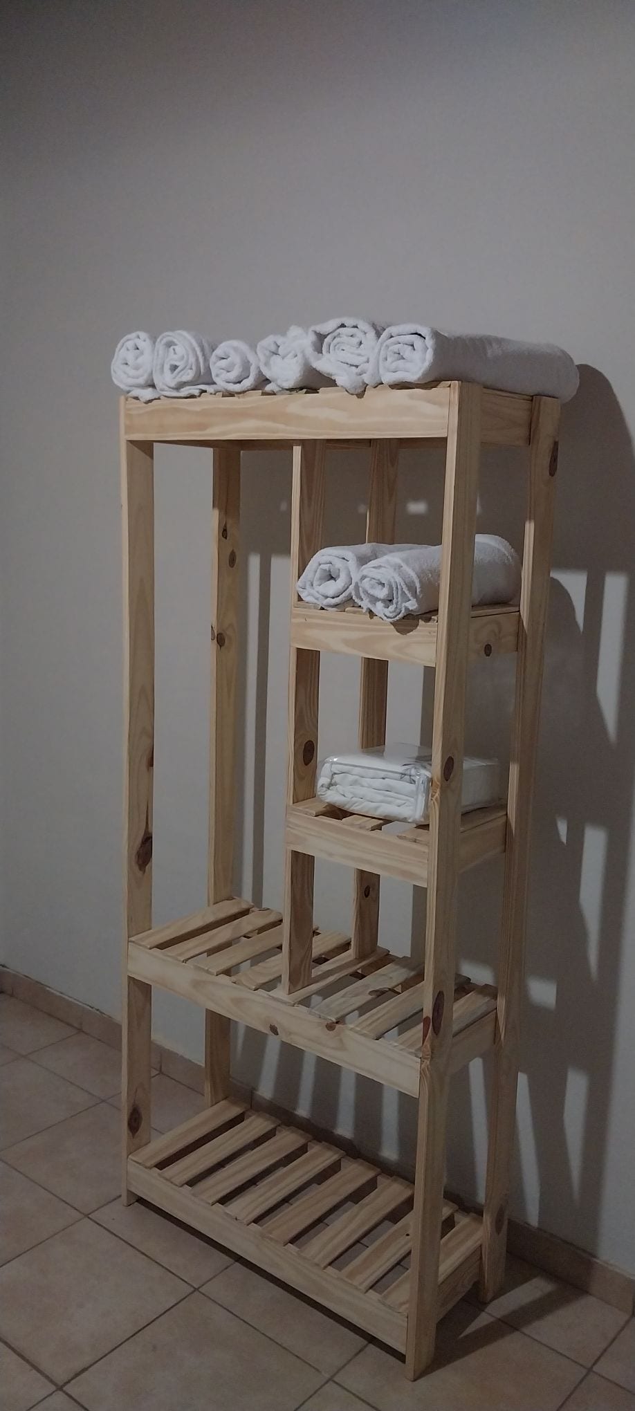 A wooden shelf is seen against a neutral wall, featuring neatly rolled white towels on multiple tiers. The shelf design incorporates open spaces for storage, with the tiles below providing a subtle texture to the setting.