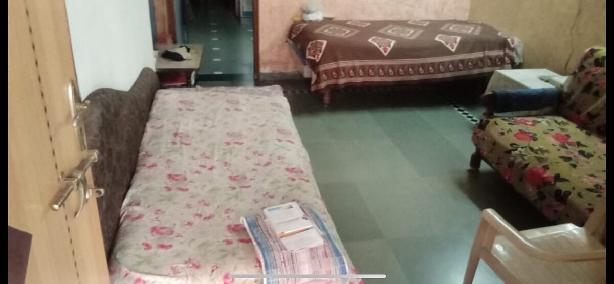 A living area is presented with two beds, one covered with a patterned blanket and the other with a floral bedspread. A wooden chair and a small table, holding some items, are visible. The flooring appears tiled and the walls are lightly colored.