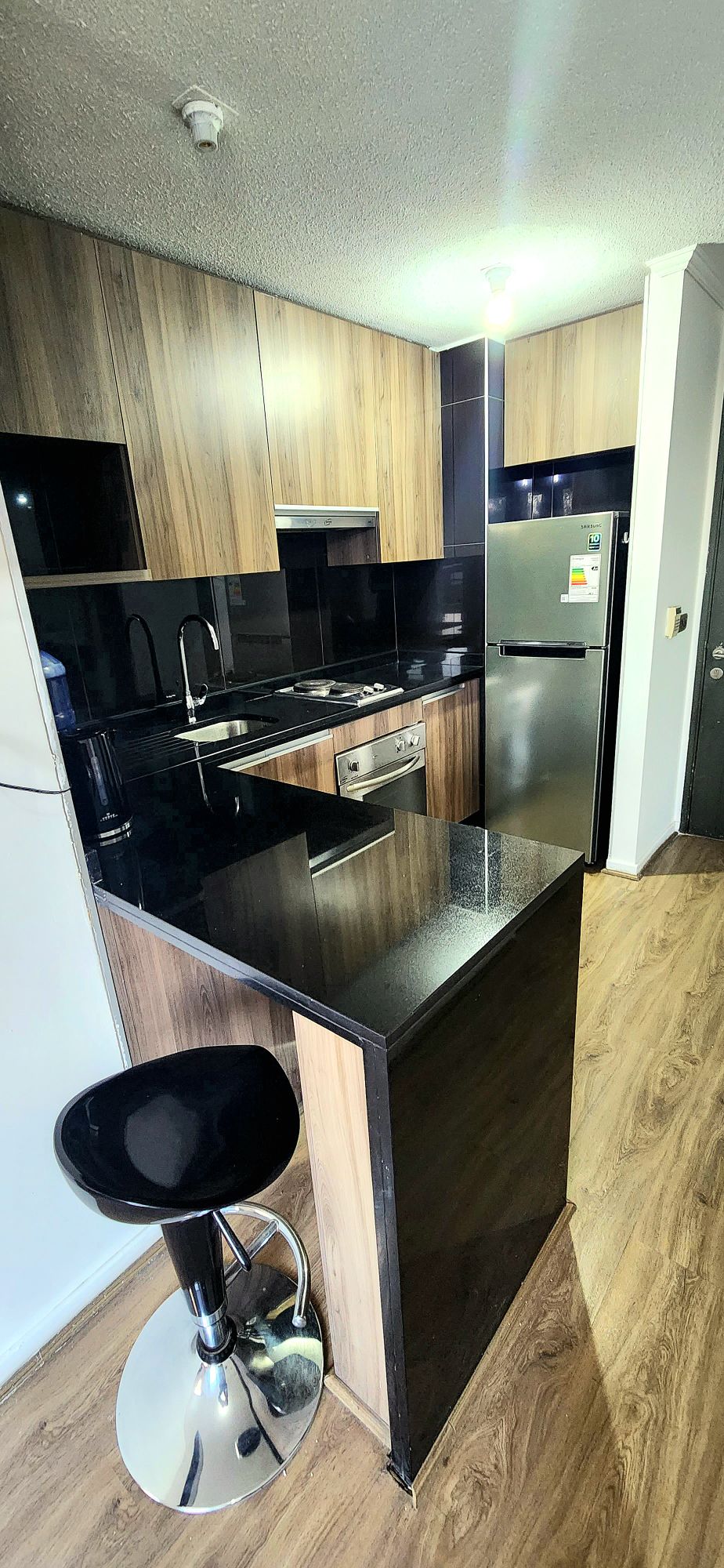 A modern kitchen is shown, featuring sleek black countertops and wooden cabinetry. Stainless steel appliances, including a refrigerator, oven, and stove, are integrated seamlessly. A barstool sits beside a raised countertop, creating a stylish dining area. Natural light illuminates the space.