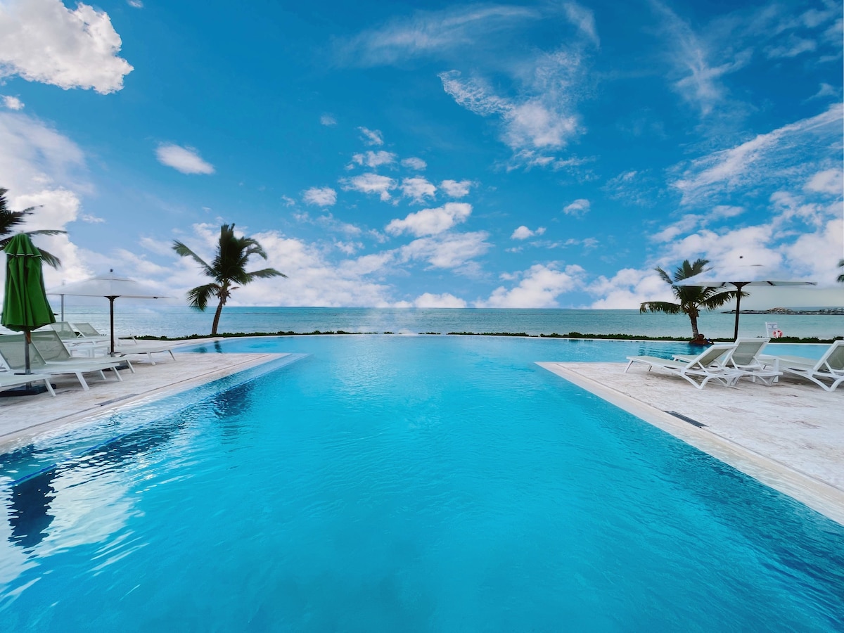 An infinity pool stretches toward the horizon, meeting the ocean under a bright blue sky filled with scattered clouds. Palm trees are visible along the pool's edge, enhancing the serene waterfront view.