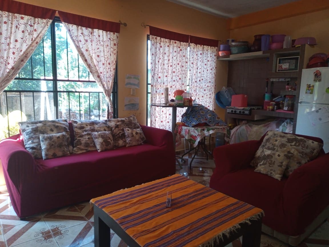 A living area is presented with two red sofas adorned with patterned cushions. A coffee table with a colorful tablecloth is centrally located. Natural light filters through floral curtains, while a kitchen area is visible in the background, containing various items and appliances.