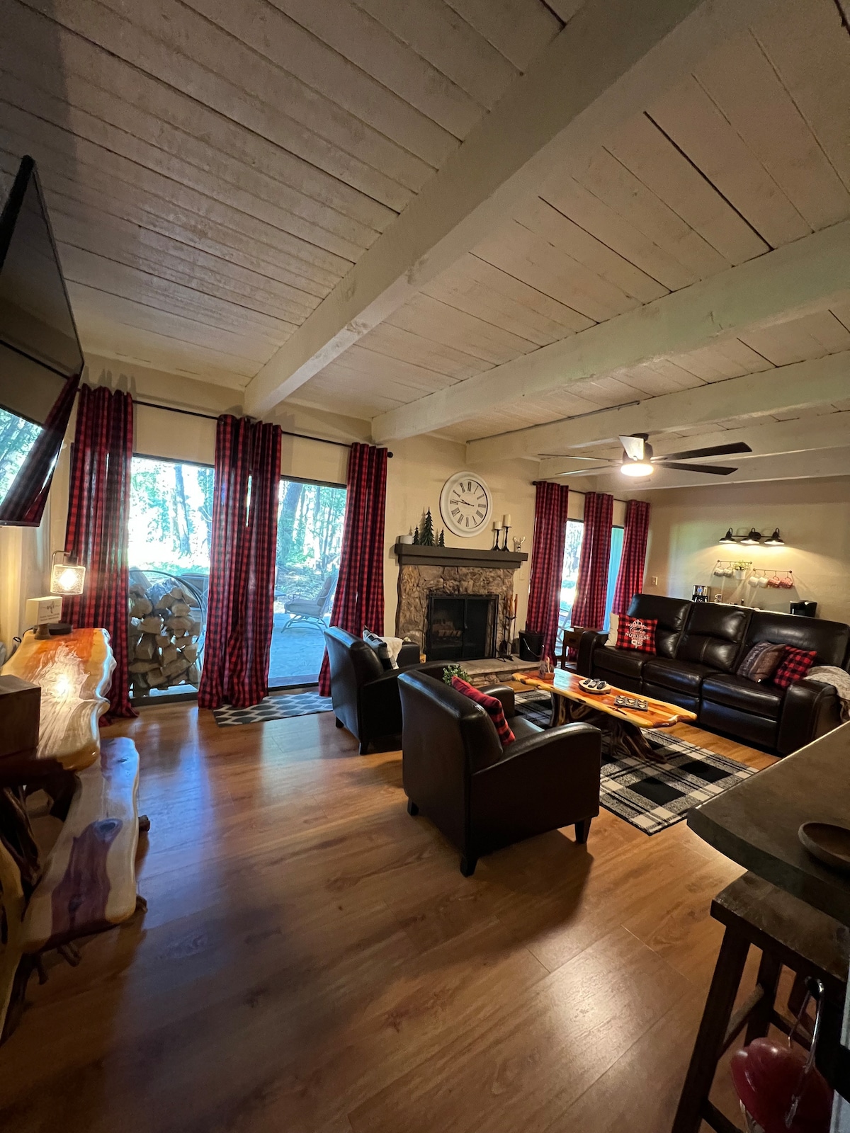 A spacious living room is highlighted by plush leather seating and a decorative clock above the fireplace. Large windows with red checked curtains allow natural light, while a rustic wood pile adds to the cozy ambiance. A patterned rug complements the wooden floors.