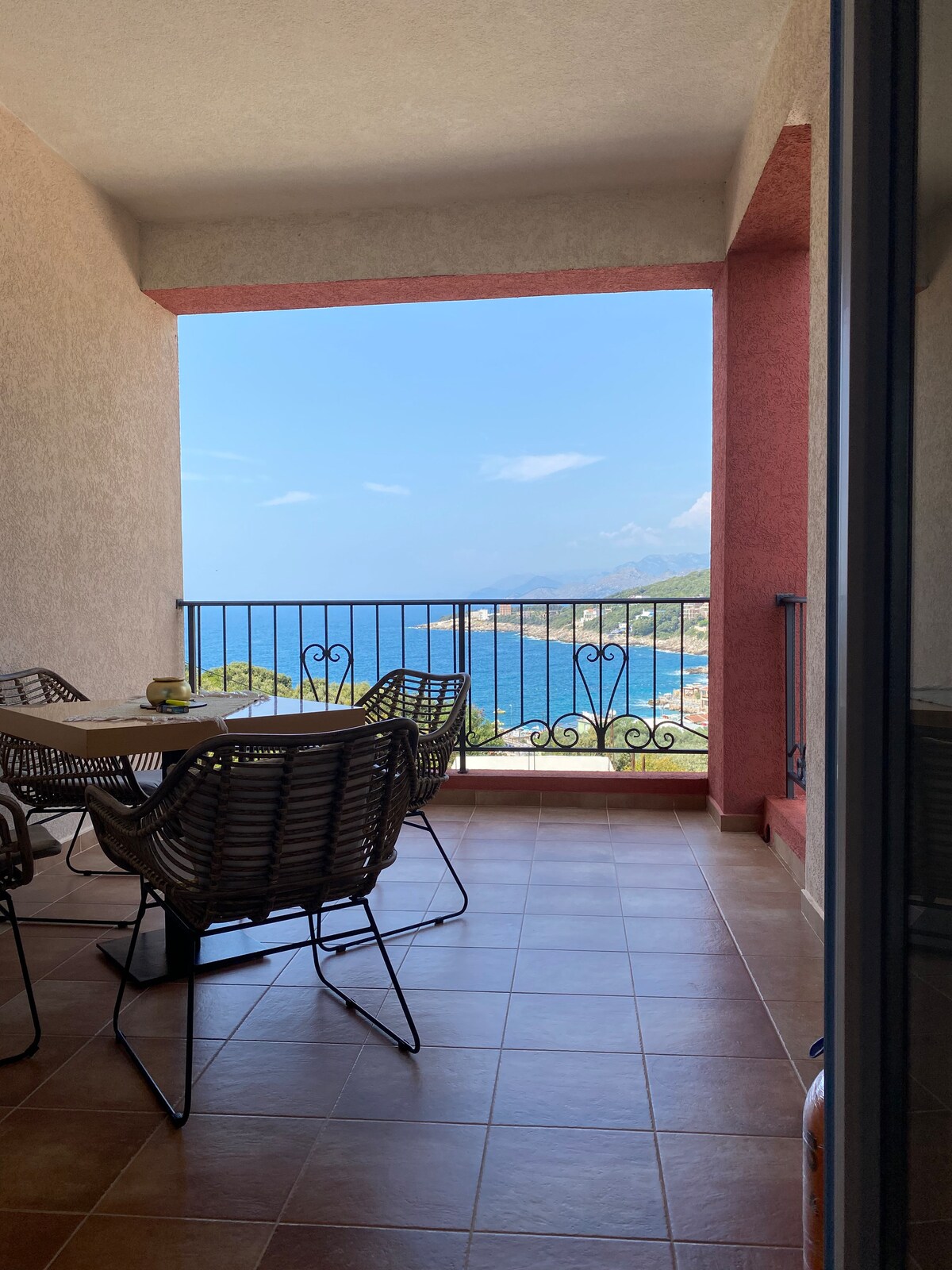 A balcony area features a dining table surrounded by several woven chairs. A railing provides a view of the sea and distant coastline under a clear sky. Natural light fills the space, highlighting the warm tile flooring.