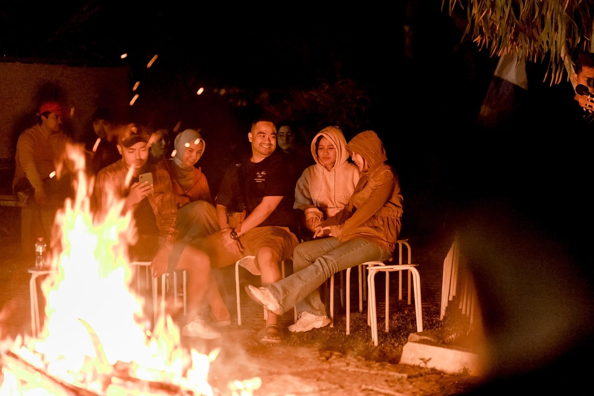 An outdoor gathering is illuminated by a warm fire at night. Several individuals are seated on white chairs, with a fire pit providing a cozy focal point. The surrounding greenery and night sky create a serene backdrop, enhancing the relaxed atmosphere.
