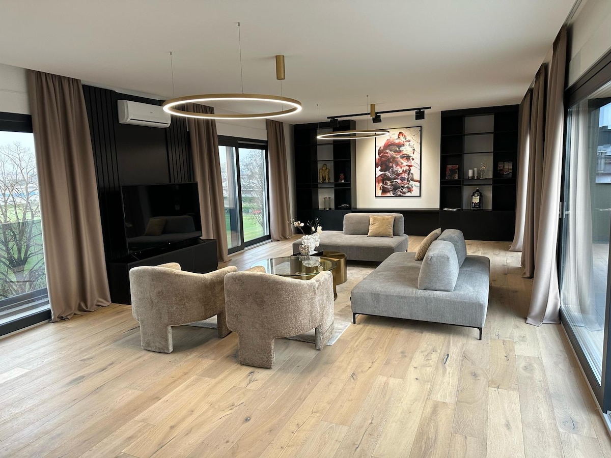 A modern living area features a sectional sofa and plush armchairs arranged around a glass coffee table. Large windows allow natural light to fill the space, while elegant curtains frame the views. A minimalist shelving unit displays artistic decor, enhancing the contemporary design.