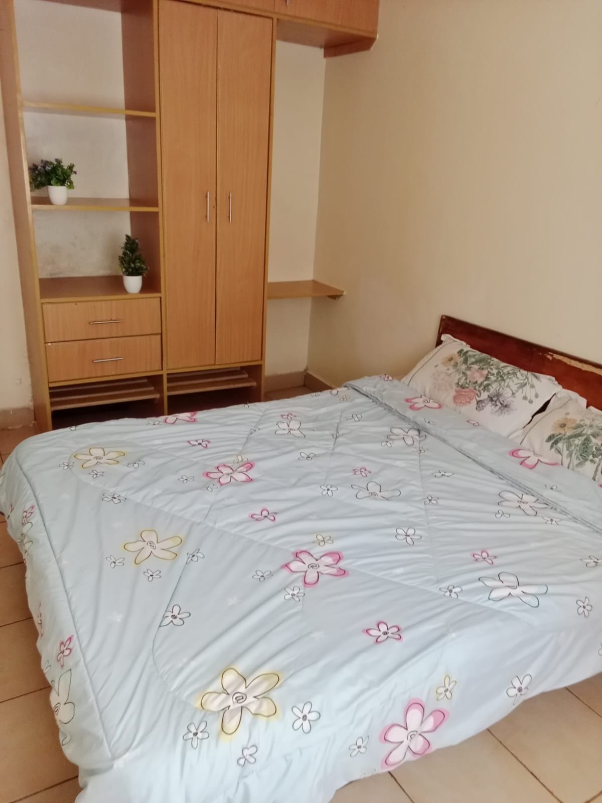 A cozy sleeping area features a spacious bed with a light blue quilt adorned with floral patterns. A wooden headboard complements the bedding. Nearby, a simple wooden closet with open shelving and drawers is visible, alongside small potted plants adding a touch of greenery.
