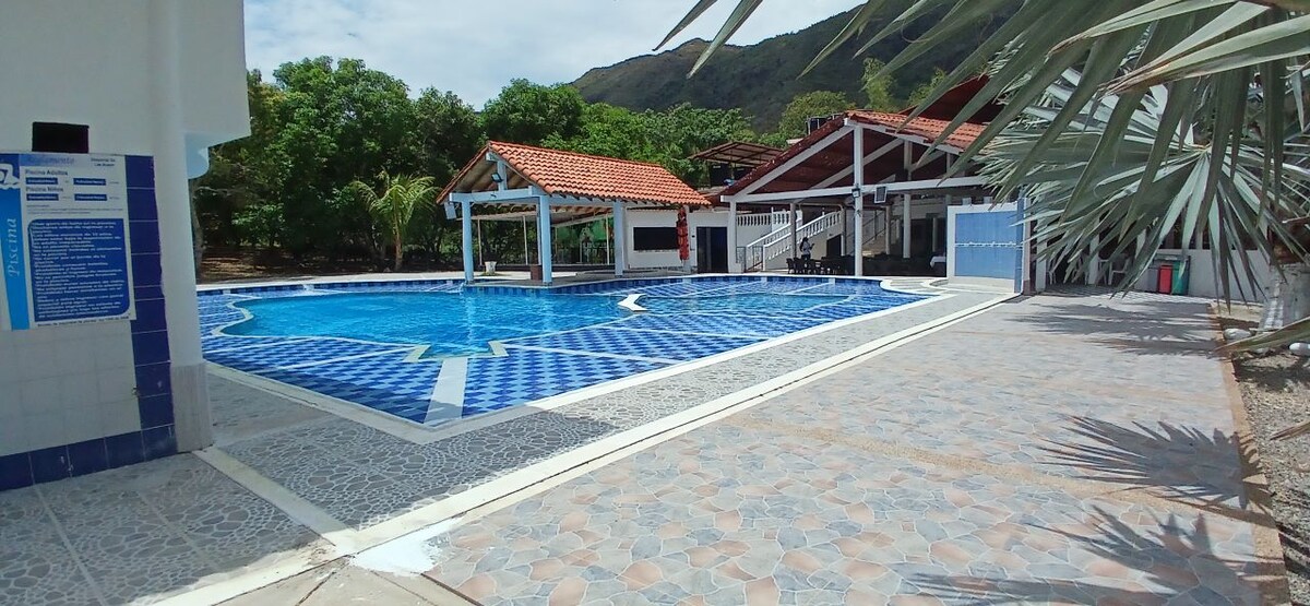 A large swimming pool is surrounded by patterned tiles, reflecting sunlight from the clear sky. Nearby, a shaded seating area and a covered pavilion can be seen, with lush greenery and distant mountains enhancing the backdrop.