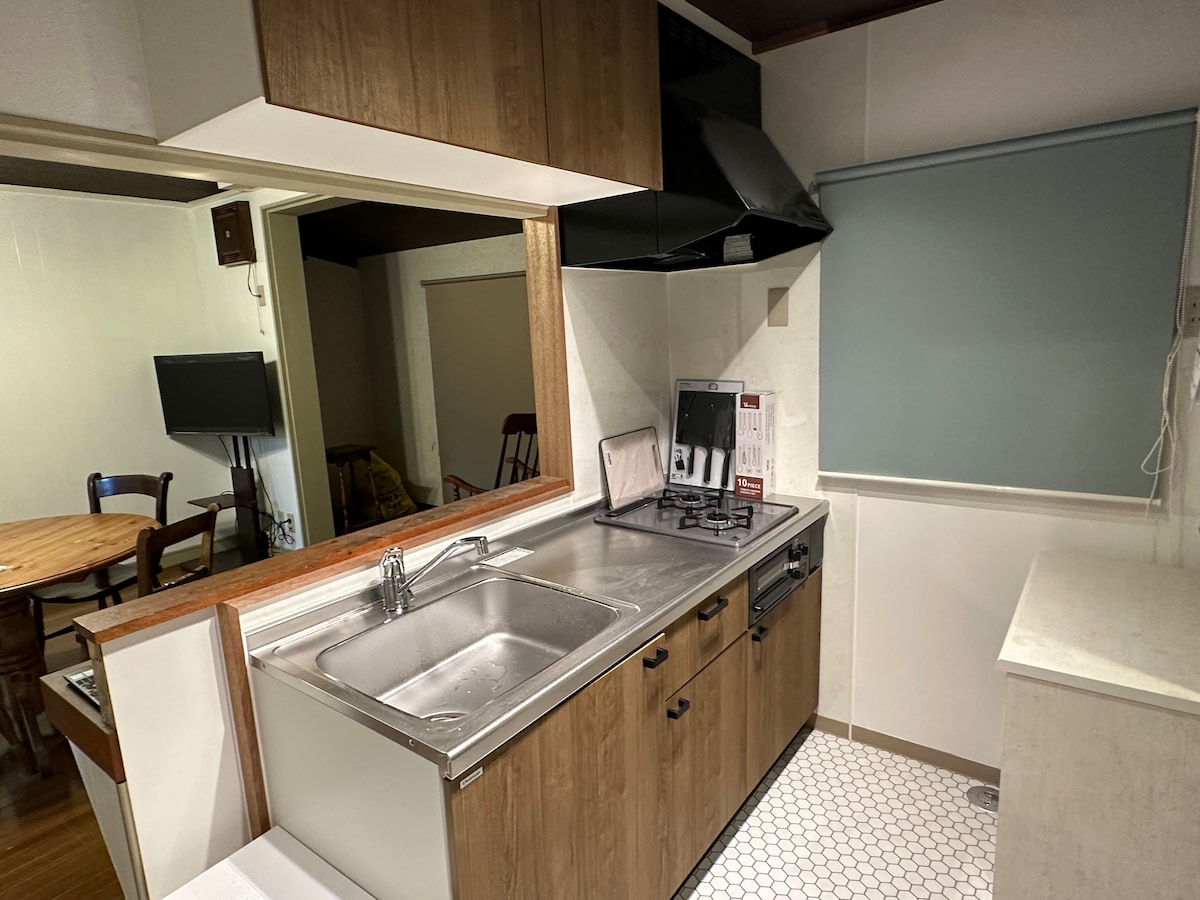 A functional kitchen space is presented, featuring a stainless steel sink and a modern gas stove. Wood cabinets and a light countertop create a warm atmosphere. A mirror reflects the dining area, which includes a wooden table and chairs, enhancing the sense of openness in the kitchen.