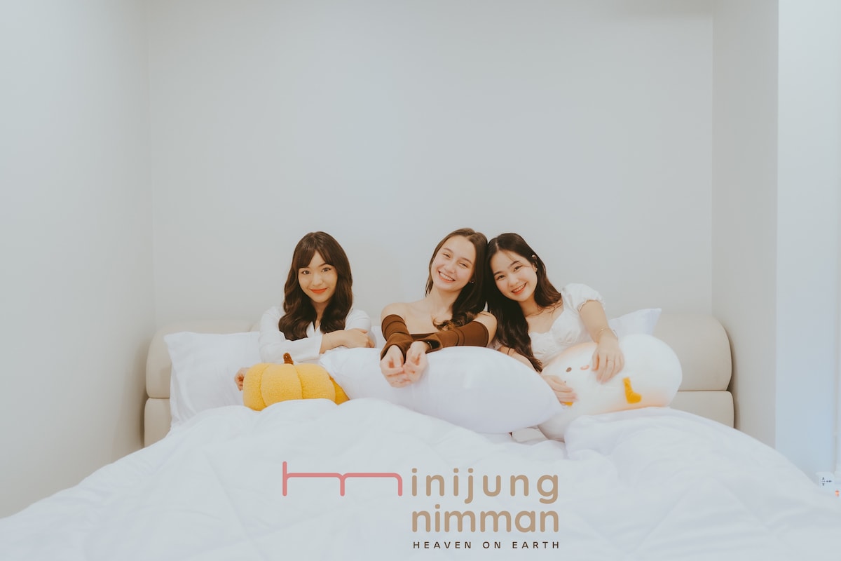A spacious bed is adorned with a white duvet and multiple decorative pillows. Three individuals are seated comfortably, surrounded by plush toys, contributing to a cozy and inviting atmosphere. Soft lighting enhances the serene space, while minimalistic décor complements the overall aesthetic.