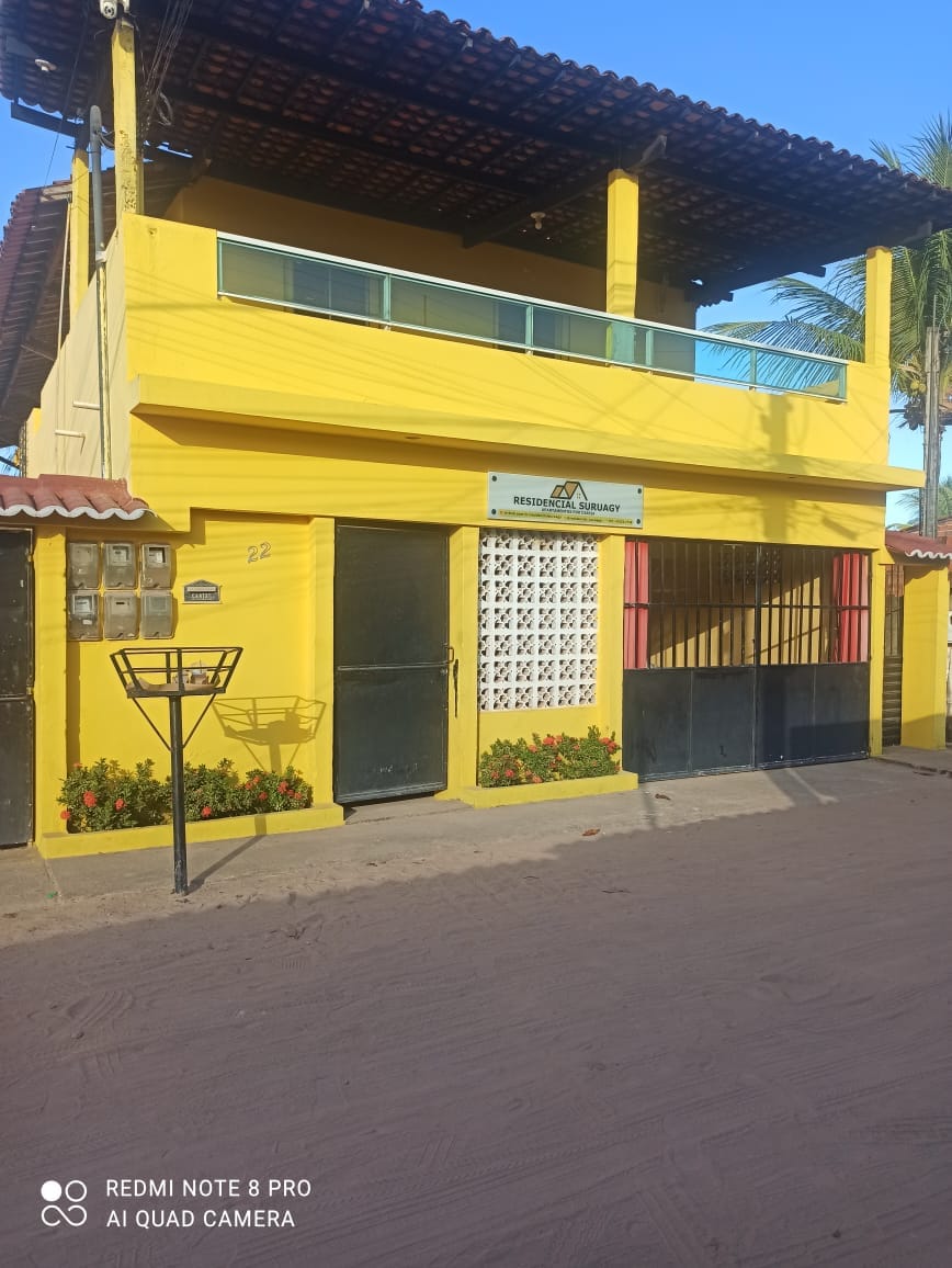 The bright yellow exterior of the property features a welcoming entrance with a gated door and ample outdoor space. A balcony with wooden railings is visible above, surrounded by greenery and tropical plants, enhancing the charm of the building.