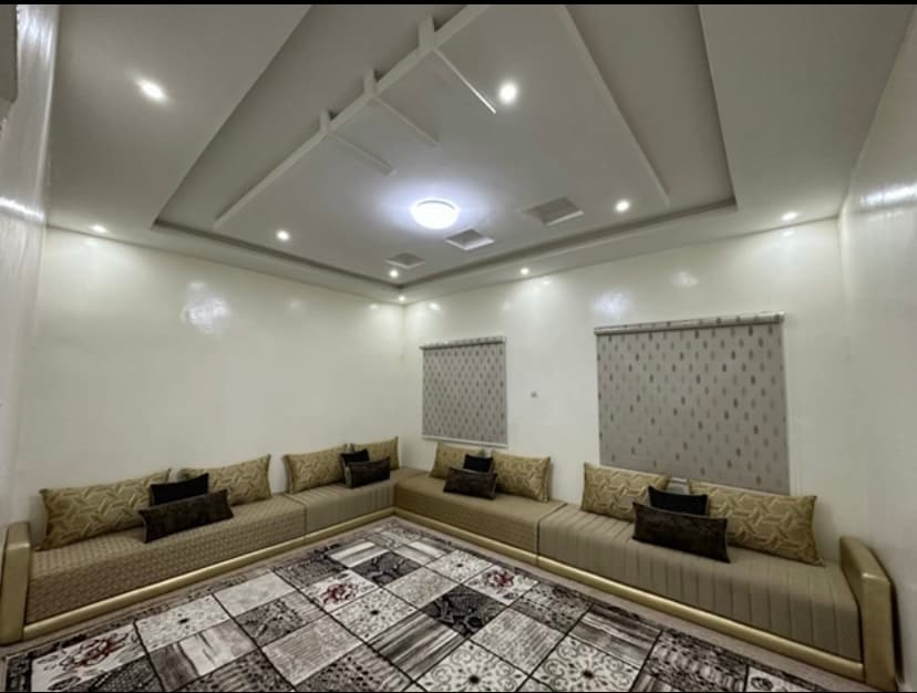 A spacious lounge area features a large, intricately patterned rug covering the floor. L-shaped seating is surrounded by decorative cushions, enhancing the relaxing atmosphere. Two windows are adorned with light filtering shades, allowing soft illumination from overhead lighting.