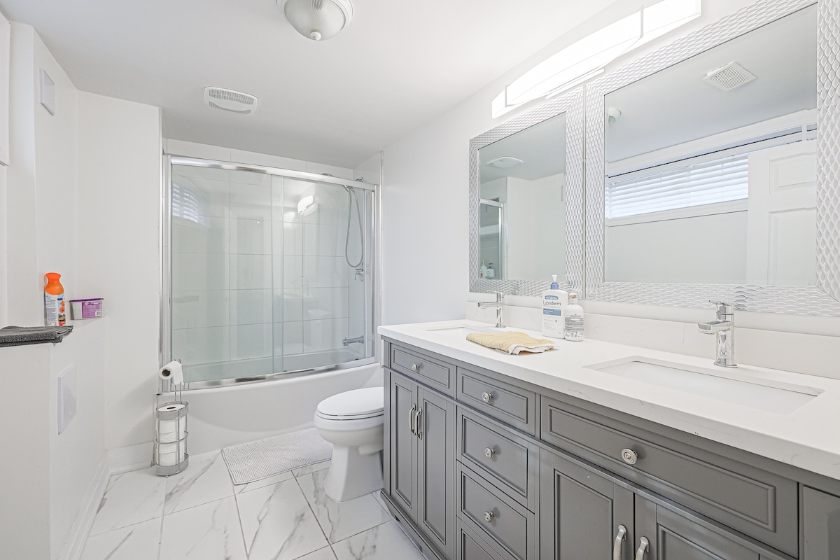 A well-appointed bathroom is equipped with dual sinks set in a modern vanity beneath large mirrors. Natural light enters through a nearby window, and a shower with glass doors complements the tub area. Neutral tiles cover the floor, creating a clean and spacious environment.