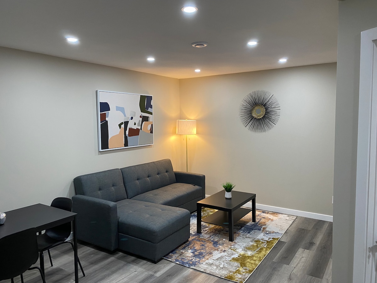 A cozy living area features a grey sectional sofa positioned beside a black coffee table. A decorative wall piece and a lamp add warmth to the space, while a colorful area rug creates an inviting atmosphere. Soft lighting highlights the modern, neutral decor.