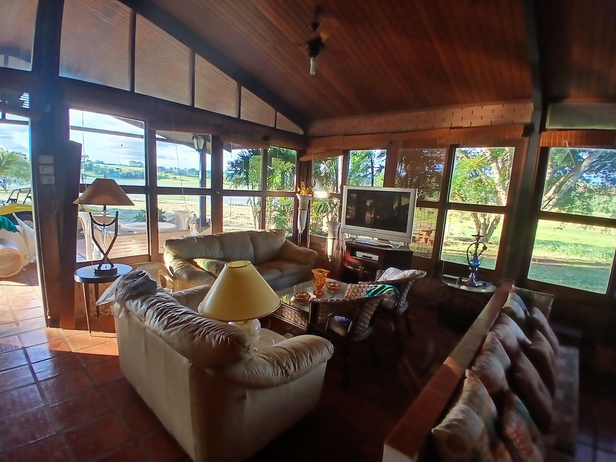 A spacious living area is surrounded by large windows, allowing natural light to fill the room. Comfortable seating is arranged around a central coffee table, and a television is positioned for viewing. A lamp provides additional lighting in the warm, wooden space.