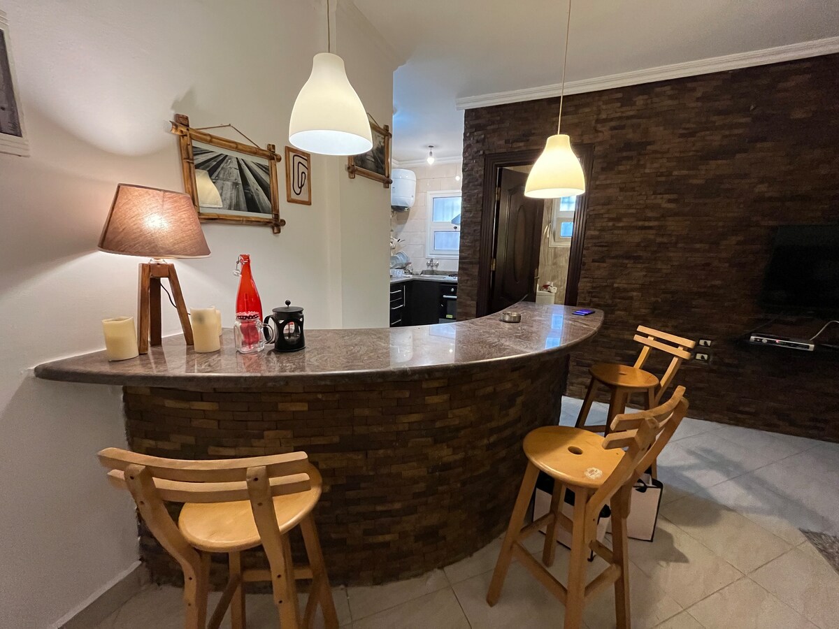 A well-appointed kitchen bar features a curved countertop made of stone, complemented by three wooden stools. Stylish pendant lights hang above, providing soft illumination. The space is accented by warm tones from the decor, enhancing the welcoming ambiance.