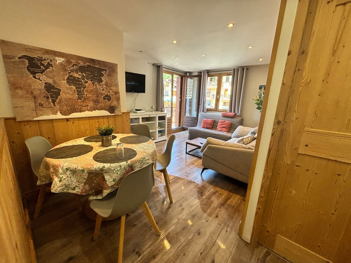 A welcoming living area features a circular dining table surrounded by four chairs, offering a cozy spot for meals. A comfortable sofa with colorful cushions faces a large window that invites natural light, while a wall-mounted TV is visible for entertainment.